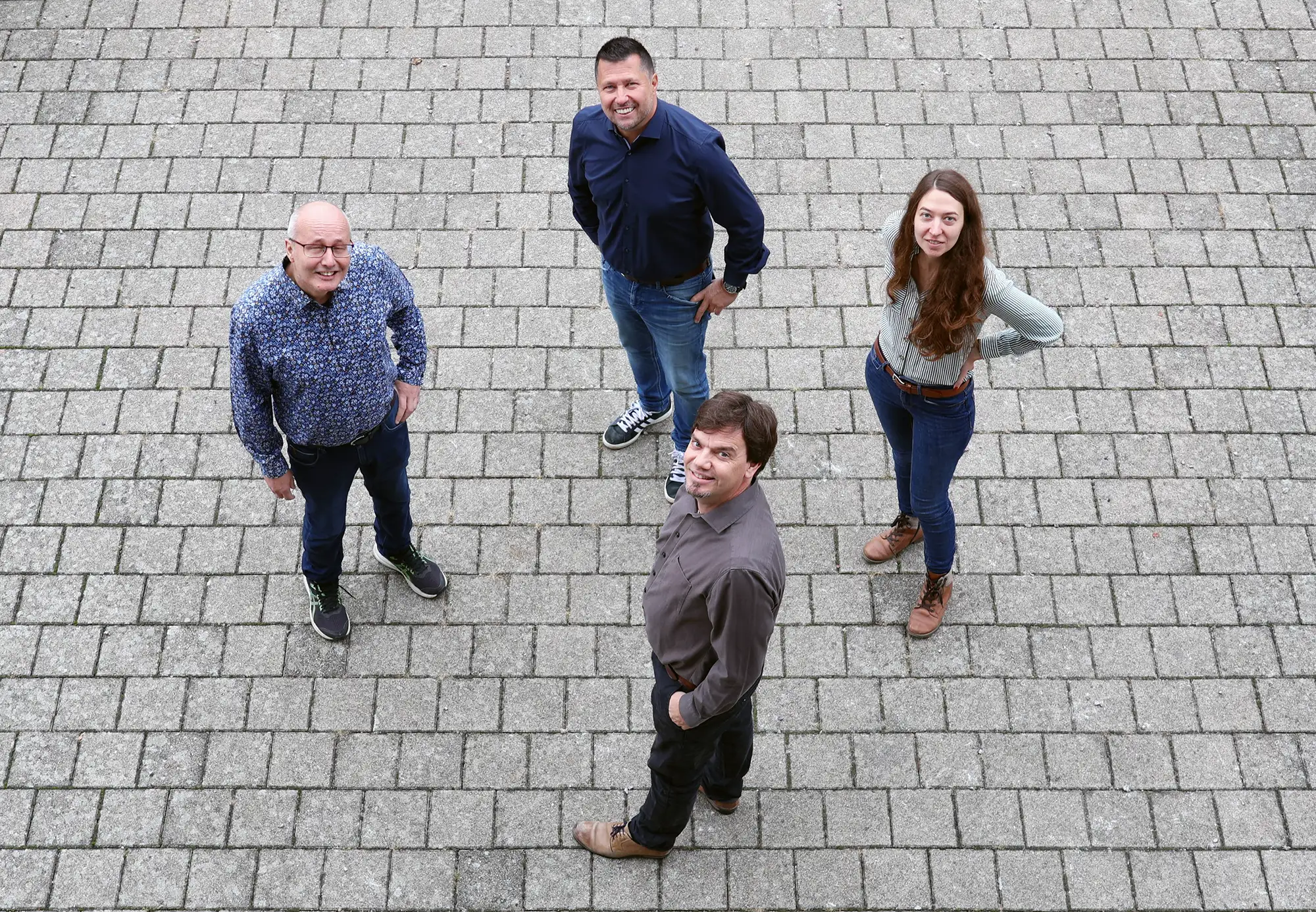Vier Personen stehen im Freien auf einem gepflasterten Platz und blicken lächelnd nach oben in die Kamera – Teamfoto von oben aufgenommen.