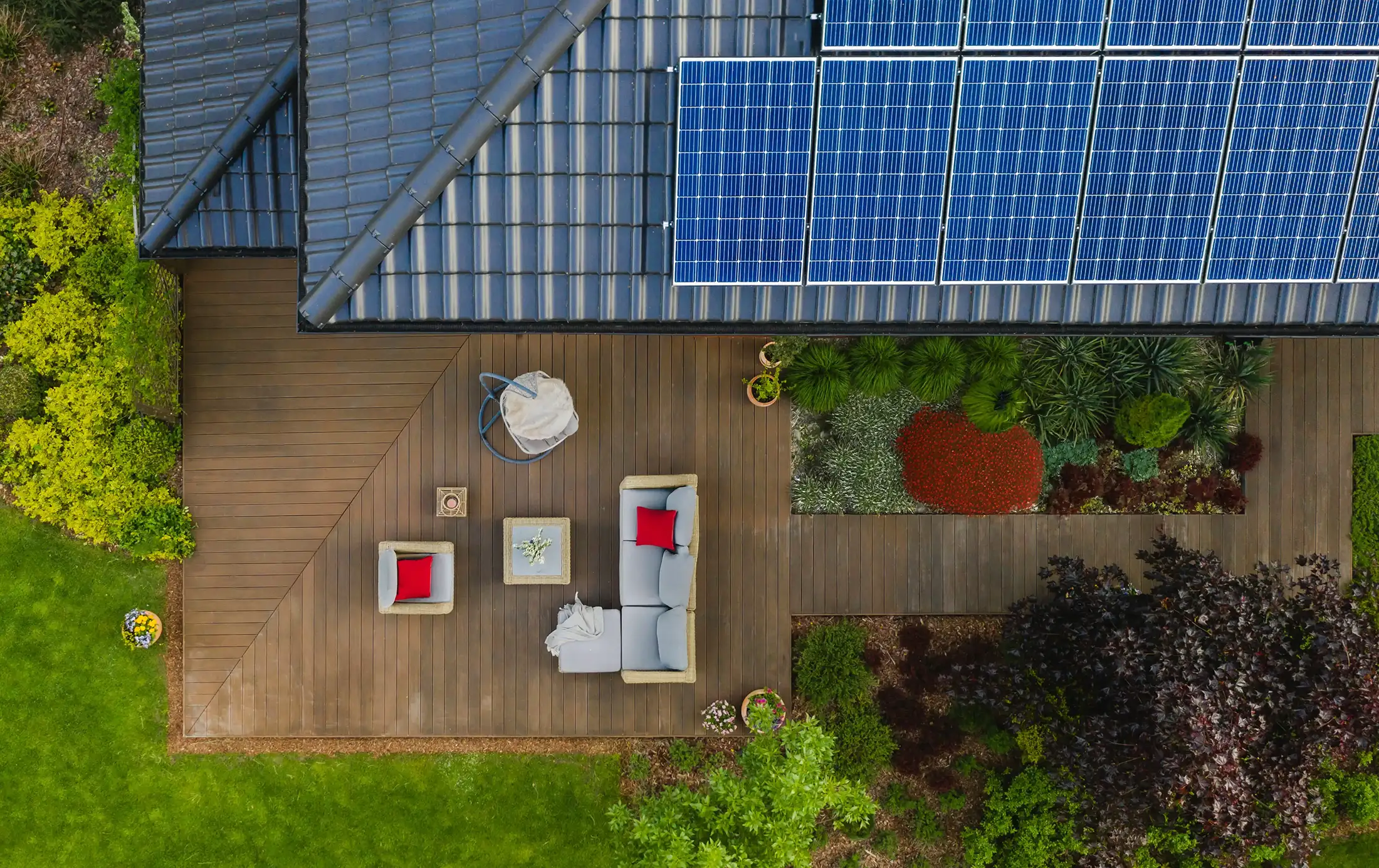 Draufsicht auf ein Hausdach mit Solarpanels und angrenzender Holzterrasse mit Sitzmöbeln, umgeben von gepflegtem Garten.