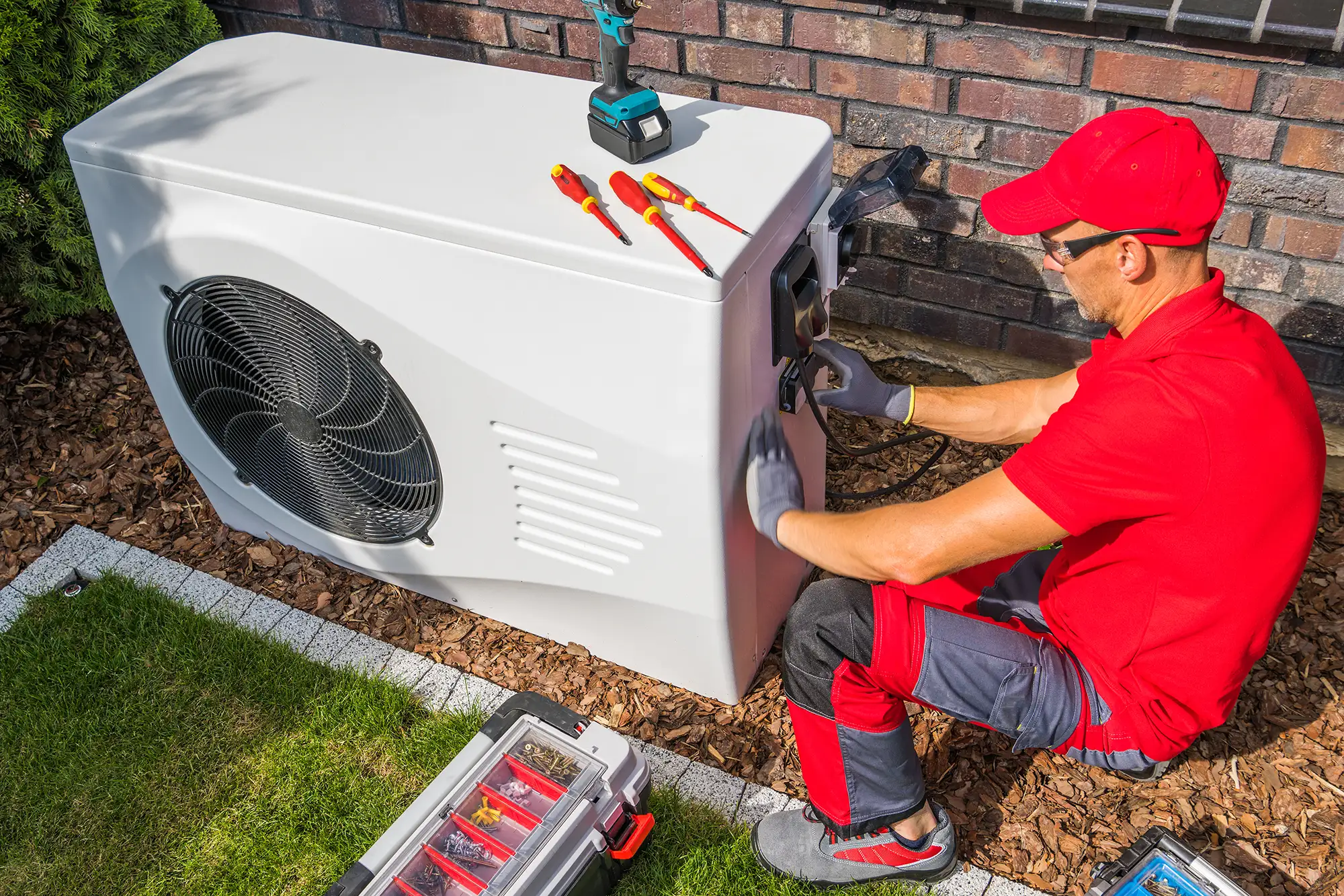 Techniker in roter Arbeitskleidung installiert oder wartet eine Wärmepumpe im Außenbereich eines Hauses.